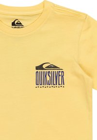 Gul t-shirt i bomull, med en svart och lila Quiksilver-logga på vänster bröstområde och geometriska accenter nedanför.
