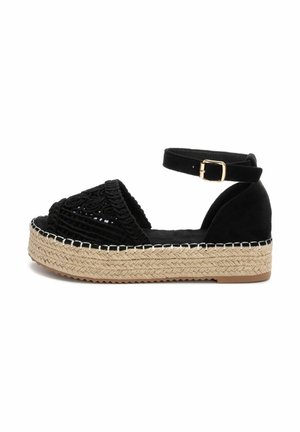Sandalia espadrille negra con parte superior de tejido trenzado, correa en el tobillo y plataforma de yute texturizado. Suela lisa con una ligera curva. Acento de hardware dorado.