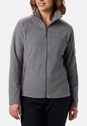 Kobieta ubrana w szarą polarową kurtkę zapinaną na zamek, z zamykanymi na zamek bocznymi i na ramionach kieszeniami, stojąca na gładkim tle.