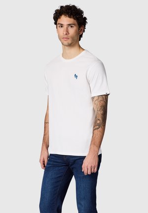Jonge man met krullend haar, draagend een effen wit T-shirt met kleine blauwe vlekken en donkerblauwe spijkerbroek, staand tegen een effen achtergrond.