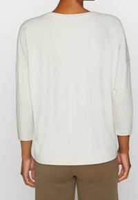 Vit topp med trekvartsärmar i mjukt tyg, med en avslappnad passform och rundad halsringning, kombinerad med bruna byxor.