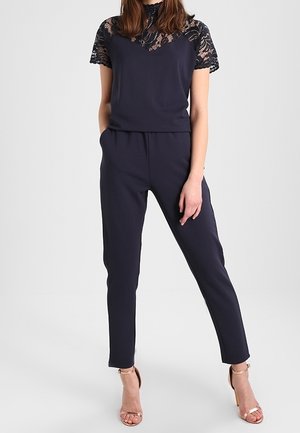 Femme portant une combinaison bleu marine avec des manches courtes en dentelle et des sandales à talons hauts dorées ouvertes à l'avant, debout devant un fond uni.