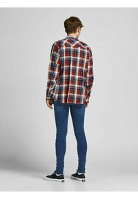 Camisa de franela a cuadros en rojo, blanco y azul marino, con mangas largas, yugo trasero y dobladillo curvado, usada sobre jeans ajustados oscuros y zapatillas negras.