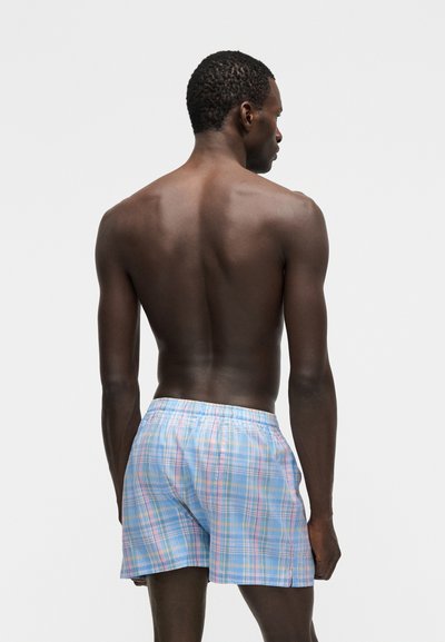 Άνδρας μοντέλο με την πλάτη, φορώντας καρό σορτς σε ανοιχτό γαλάζιο και ροζ χρώμα, με φόντο απλό ανοιχτό γκρι.