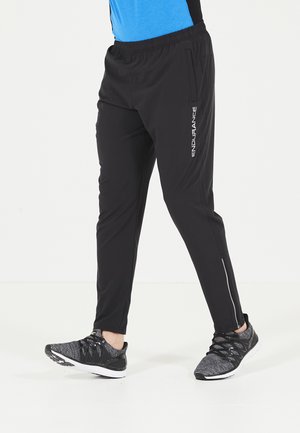 Pantalons de sport noirs à coupe fuselée, avec une taille élastique, des poches zippées et des détails de logo réfléchissant sur le côté. Portés avec des baskets noires.