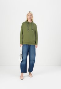 Olivnozeleni bluza s kapuco in vrvico, v kombinaciji s širokimi modrimi hlačami. Model drži majhno belo torbico in nosi svetlo rožnate balerinke.