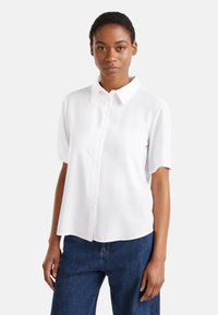 Blusa bianca a maniche corte con colletto a punta, chiusura frontale con bottoni e tessuto liscio. Abbinata a jeans blu a vita alta.