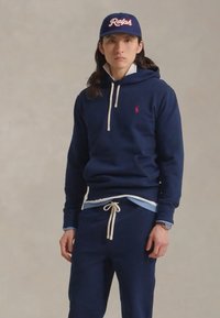 Junger Mann in navyblauem Hoodie und Jogginghose, mit blauer Mütze mit dem Text "Ralph", stehend vor schlichtem Hintergrund.