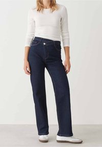 Woman wearing dark blue high-waist straight-leg jeans, white long-sleeve top, and white sneakers standing against a plain background.