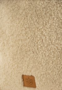 Crèmekleurige, gestructureerde stof met een zachte, pluizige textuur en een klein bruin leren label met een ster- en schildlogo.