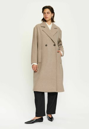 Beige Wollmischung Doppelreiher mit großen Revers, zwei Vorderaschen und Knopfdetails, getragen über schwarzen Nadelstreifen-Hosen und Ballerinas.