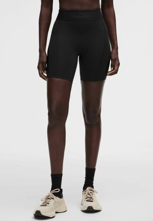 Persoon die zwarte, hooggetailleerde sportshorts, zwarte sokken en beige sneakers draagt, staand tegen een effen lichtgrijze achtergrond.