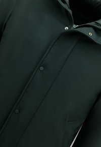 Zwarte waterdichte jas met ritssluiting, drukknoopsluiting en een hoge kraag. Heeft een gladde, matte textuur en zijzakken.