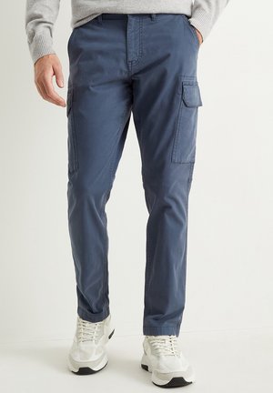 Mężczyzna w niebieskich spodniach typu cargo z bocznymi kieszeniami i białych sneakersach, stojący z jedną ręką w kieszeni na jednolitym jasnym tle.