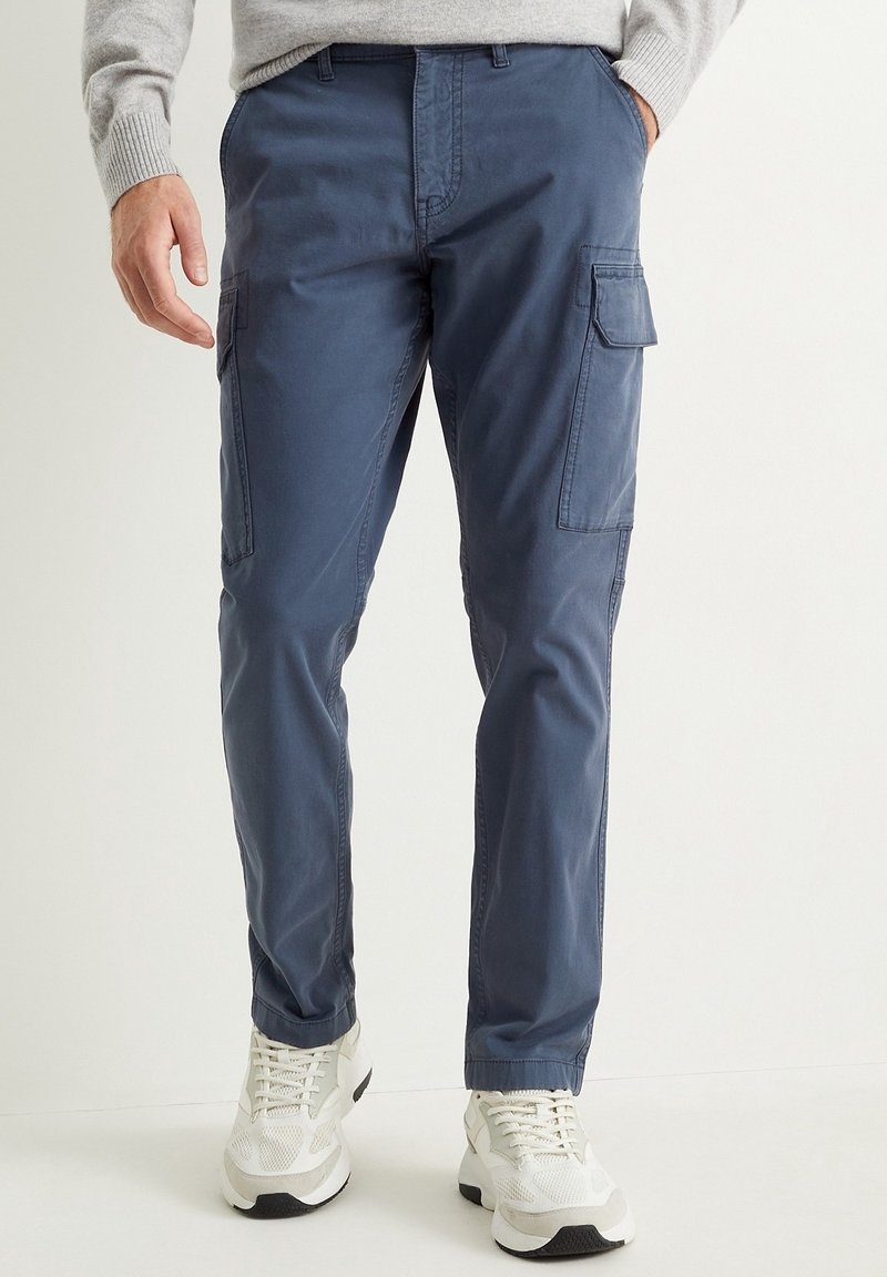 Homme portant un pantalon cargo bleu avec des poches latérales et des baskets blanches, debout avec une main dans la poche devant un fond clair uni.