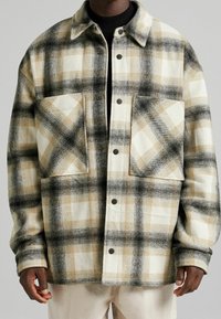 Hombre vistiendo una chaqueta de cuadros beige, negra y blanca con dos grandes bolsillos en el pecho sobre un cuello de tortuga negro.