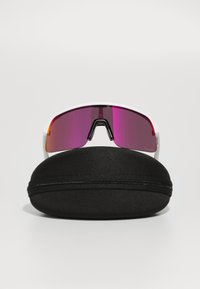 Gafas de sol deportivas con montura blanca y lentes rosa espejo, guardadas en un estuche rígido redondo negro con cierre de cremallera.