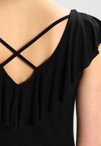 Zadní pohled na osobu v černých šatech s volánovými rukávy a tenkými křížovými ramínky na horní části zad.