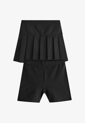Jong meisje in witte blouse en zwarte rok speelt met een houten jojo naast een houten stoel op een lichte achtergrond.