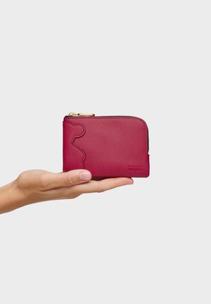 Mano sosteniendo una pequeña cartera roja con textura y cremallera dorada, con un diseño en relieve de piezas de rompecabezas en el lateral.