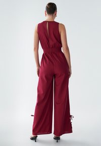 Bordo kombinezon brez rokavov, z elastičnim pasom, odprtim hrbtom in širokimi hlačnicami z vezalkami na gležnjih za prilagajanje.