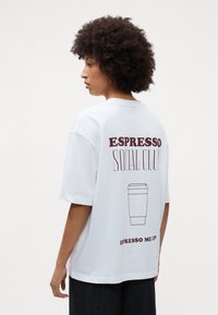 Balta medvilninė marškinėliai su per dideliu kirpimu, su bordo spalvos tekstu "ESPRESSO SOCIAL CLUB" ir kavos puodelio grafika ant nugaros. Trumpi rankovės.