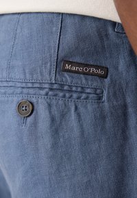 Shorts din in ușor albastru au un buzunar posterior cu o etichetă din material negruinscrisă "Marc O'Polo" și un nasture închis la culoare cu detalii ale logo-ului.