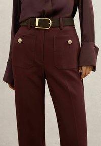 Burgundy trousers made of smooth fabric, featuring a black belt with a gold buckle and decorative buttons on each pocket.