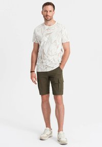 Tenue masculine composée d'un t-shirt blanc à motif floral et de shorts cargo vert olive, associés à des baskets blanches et beiges.