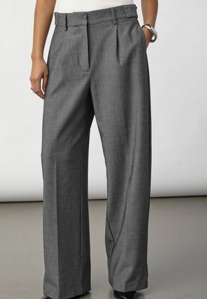 Personne portant un pantalon gris à plis, taille haute et jambes larges, mains partiellement dans les poches, debout sur un sol réfléchissant.