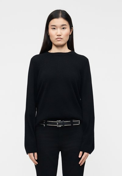 Jeune femme aux longs cheveux noirs portant un pull noir, un pantalon noir et une ceinture noire cloutée, debout devant un fond blanc.