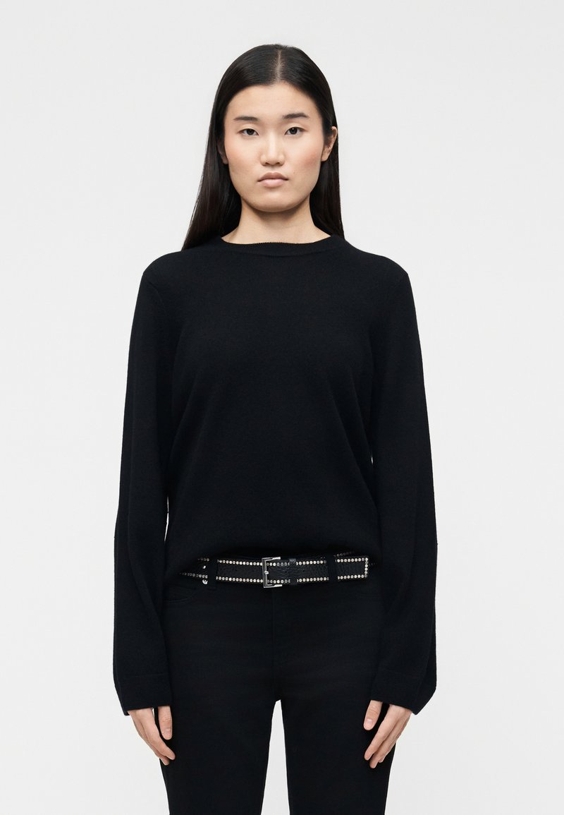 Νέα γυναίκα με μακριά σκούρα μαλλιά, φορώντας μαύρο πουλόβερ, μαύρο παντελόνι και μαύρη ζώνη με τρουκς, στέκεται μπροστά σε λευκό φόντο.