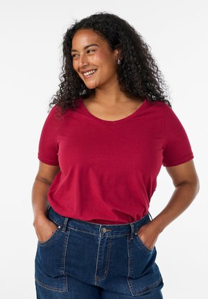 Kvinde med krøllet hår iført en rød kortærmet skjorte og blå jeans, smilende og kigger til siden med hænderne i lommerne.