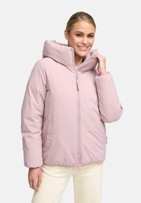 Gladde vrouw met een lichte roze gewatteerde jas met hoge kraag en rits, gecombineerd met crèmekleurige broek, staande tegen een witte achtergrond.
