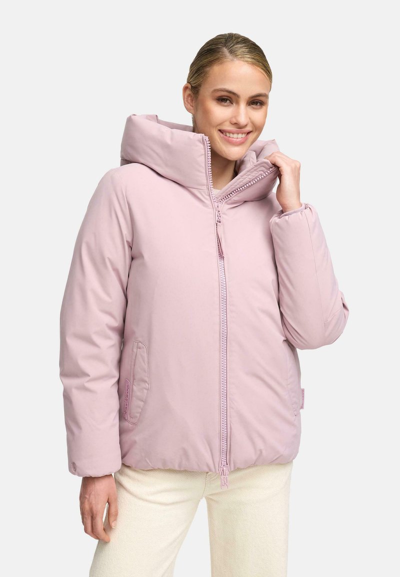 Gladde vrouw met een lichte roze gewatteerde jas met hoge kraag en rits, gecombineerd met crèmekleurige broek, staande tegen een witte achtergrond.