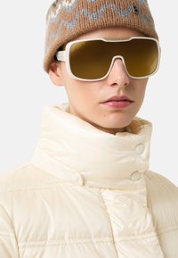 Białe, oversize okulary przeciwsłoneczne z brązowymi szkłami, w połączeniu z kremową kurtką puchową i wzorzystą brązową czapką z dzianiny.