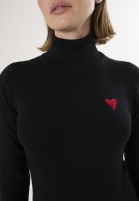 Zwarte coltrui gemaakt van zachte brei-stof, met een klein rood hart geborduurd op de linkerkant van de borst.