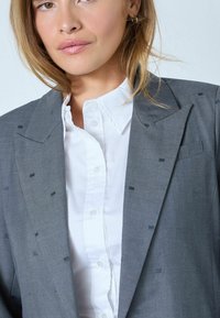 Blazer gris con un sutil patrón texturizado y solapas con muesca, llevado sobre una camisa blanca de botones con un cuello nítido.