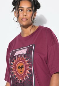 Frau mit dunklem Haar in lockeren Duttfrisuren trägt große Kreolen und ein kastanienbraunes T-Shirt mit einem stilisierten Sonnen-Gesicht und der Zahl 3.