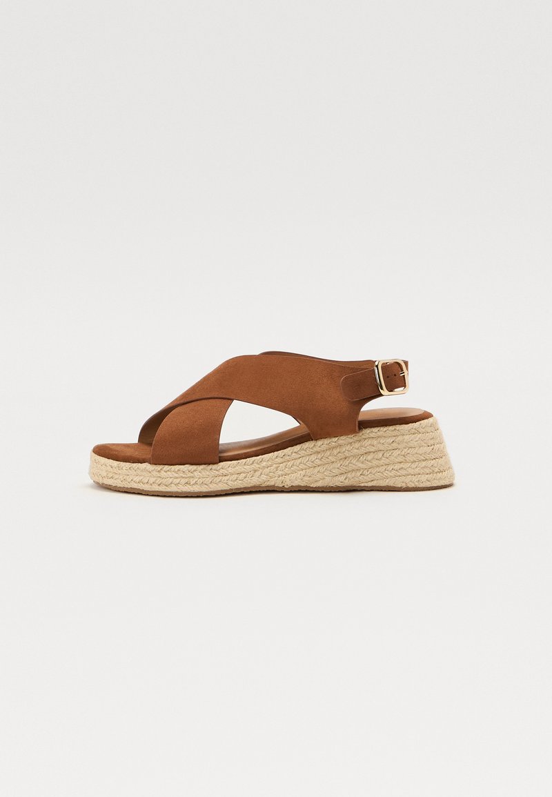 Brown suede wedge sandals featuring crisscross straps, a woven jute platform, and an adjustable ankle strap with a gold buckle accent.