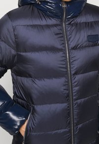 Marinblå pufferjacka med en glänsande huva och dragkedjestängning. Har horisontell quiltad design och sidofickor. Släta, glänsande ärmar.