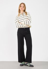 Melni plaši bikses ar pogu, kombinētas ar krēmkrāsas svītrainu garām piedurknēm tīruli un tumšām sporta kurpēm ar zaļiem akcentiem.