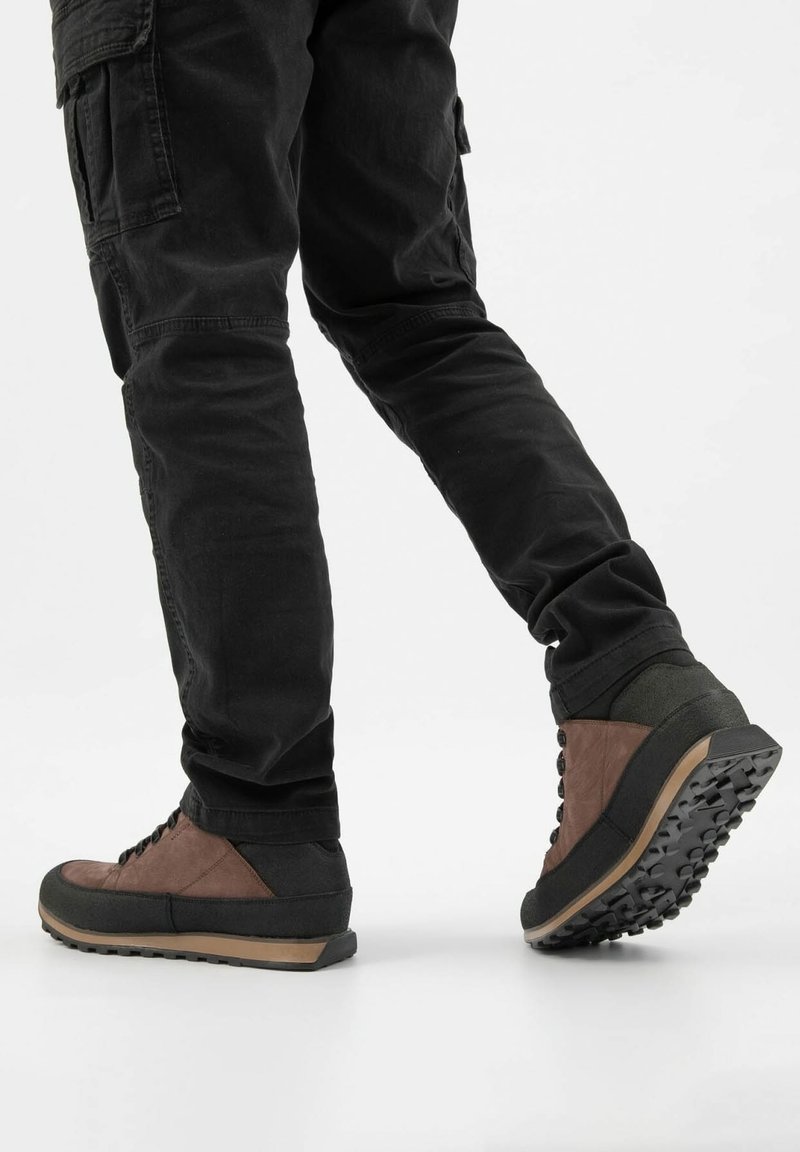 Personne portant un pantalon noir et des chaussures de randonnée marron et noires à semelles texturées, posant le pied sur une surface blanche, vue de côté et de dos.