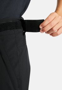 Čierne nohavice majú nastaviteľný pás na suchý zips, sú vyrobené z pevného materiálu a majú štruktúrovaný dizajn, vrátane bočných vreciek a upravených švov.