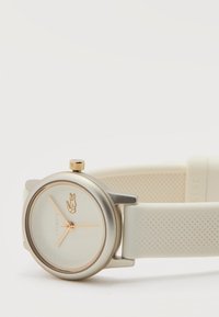 Στρογγυλό ασημένιο και χρυσό ρολόι χειρός με κρεμί υφασμένο λουράκι σιλικόνης, χρυσά δείκτες και χρυσό λογότυπο κροκόδειλου στο καντράν.