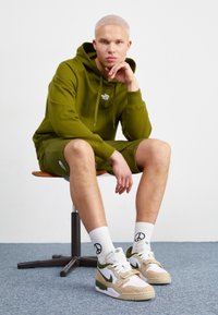 Olive green hoodie and shorts, white sneakers with accents, textured tan pattern, and white socks featuring a peace symbol, seated on a chair.