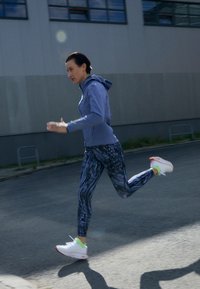 Vaaleanlila vetoketjuhuppari, kuviolliset tummat leggingsit ja valkoiset lenkkarit neonkorostuksilla. Juoksija on liikkeessä kadulla.