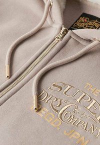 Lichtbeige hoodie met een gouden rits, reliëflogo en koorden. Zachte stof met contrasterende gouden accenten.