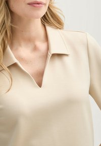 Camiseta beige de cuello en V con un diseño con cuello y textura suave, que presenta hombros sin costuras y un ajuste relajado.