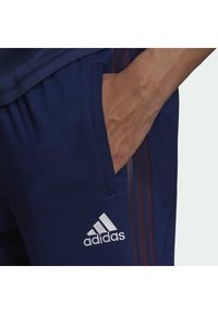 Pantaloni sportivi navy con tasca laterale, caratterizzati da tre strisce marrone scuro e un logo Adidas bianco. Tessuto morbido con una texture liscia.